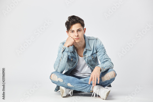 Dark-haired guy dressed in a white t-shirt, jeans and a denim jacket is sitting cross-legged on the floor on the white background in the studio