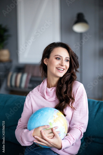 Smiling beautiful woman holding in hands a model of the globe sitting on a sofa.