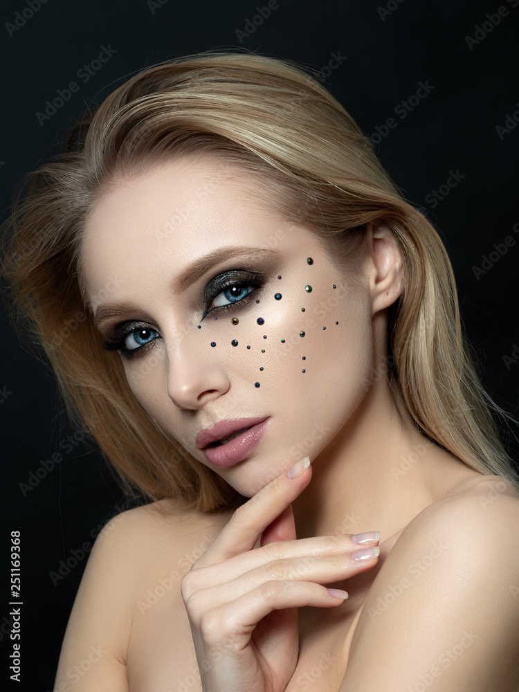 Close up beauty portrait of young woman with beautiful fashion makeup touching her face. Modern fashion makeup. Long lashes, bronze smokey eyes, black rhinestones. Studio shot. Close up beauty portrait of young woman with beautiful fashion makeup touching her face. Modern fashion makeup. Long lashes, bronze smokey eyes, black rhinestones. Studio shot.