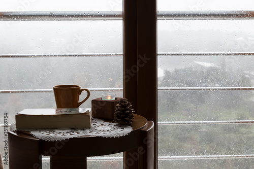 The cup of tea or coffee with a book and candle on the wooden table near the window on the rainy weather.