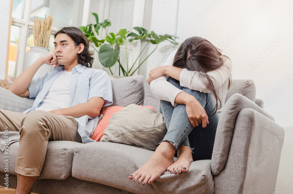 sadness young asian wife having quarrel and sitting on sofa after fight with husband behind her in house interior together, upset couple, love, divorce couple, family issues and relationship concept