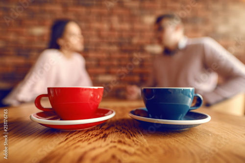 Close up of two cups of coffee on desk. In background blurred couple flirting.