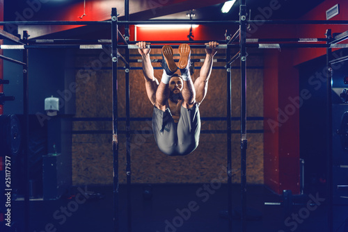 Shirtless Caucasian bearded bodybuilder doing chin ups with legs up. Gym interior, night work out concept.