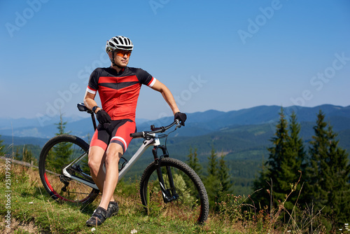 Young athletic tourist biker resting at his bike on mountain hill on bright blue summer sky and distant mountains background. Tourism, active lifestyle and extreme sport concept.