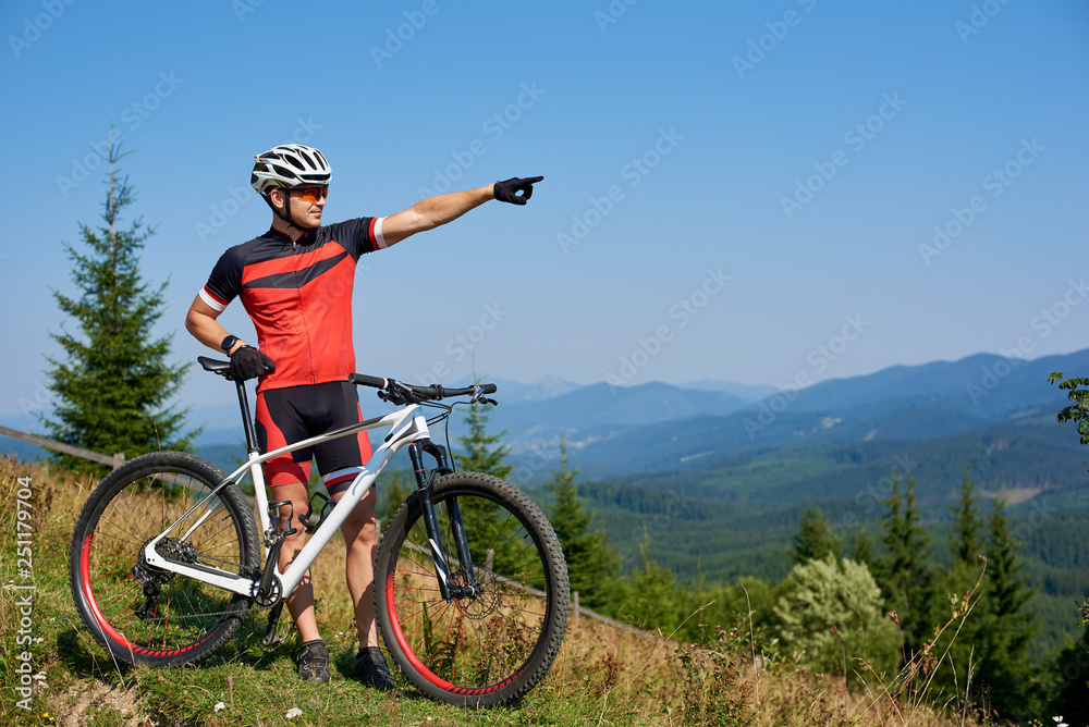 Young athletic professional biker standing with bicycle on top of hill, pointing into distance, enjoying view mountains, blue summer sky on background. Copy space. Active lifestyle and sport concept