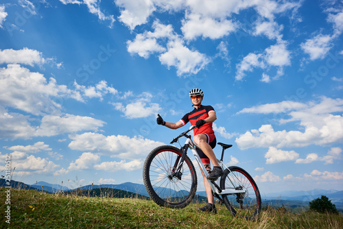 Smiling professional cyclist in sportswear and helmet standing with cross country bike on top of hill, showing thumbs up, against blue sky with clouds on summer sunny day. Outdoor sport concept
