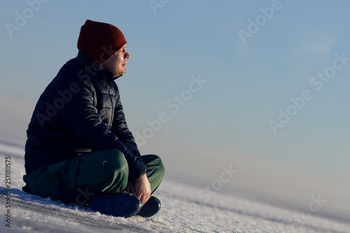 a young man sits in the winter on the ice of snow and looks at the sun.