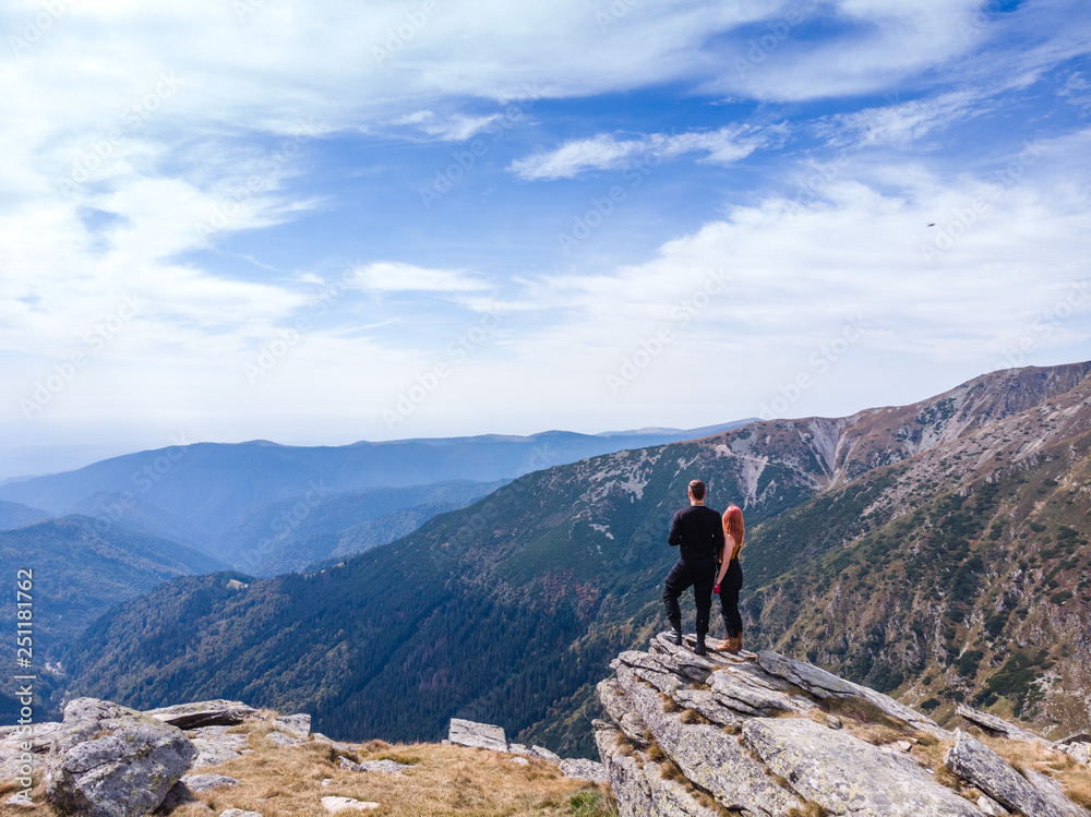 Fototapeta premium Back view of tourist couple, man and girl stand on rocky mountain top enjoying breathtaking autumn mountain panorama. Tourism, traveling and healthy lifestyle concept. motorcyclists