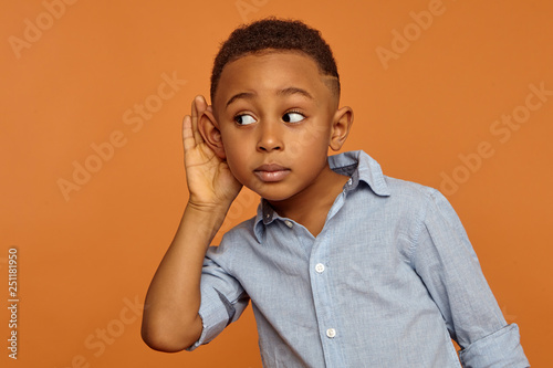Secrecy and confidential information concept. Portrait of funny curious African schoolboy eavesdropping, touching ear and looking away with surprised interested facial expression, posing in studio