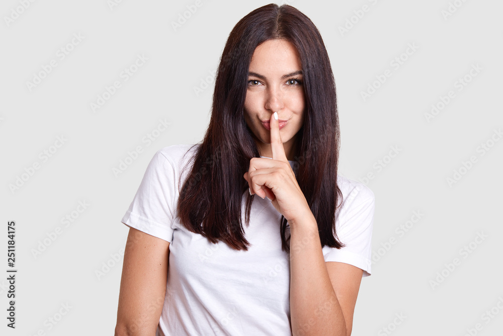 Girl like sly poses in photography. This photo is isolated. Background gray tint where model is in center. Lady wears white t shirt that fits impeccably to manicure on her finger near mouth. Girl like sly poses in photography. This photo is isolated. Background gray tint where model is in center. Lady wears white t shirt that fits impeccably to manicure on her finger near mouth.