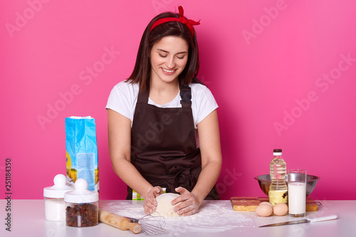 Horizontal shot of cheerful housewife kneads dough for making pizza  uses different ingredients flour  egss  oil  milk  being in good mood  wears apron  poses against pink background. Handmade