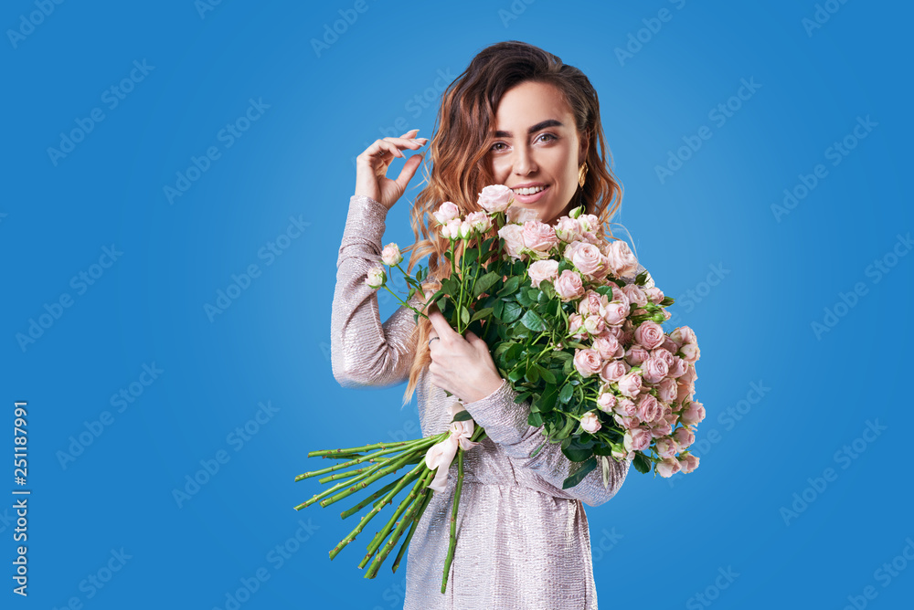Portrait of happy brunet young woman with bouquet of pink spring flowers over blue background. Festive bouquet in honor of women's day on March 8 or birthday Portrait of happy brunet young woman with bouquet of pink spring flowers over blue background. Festive bouquet in honor of women's day on March 8 or birthday