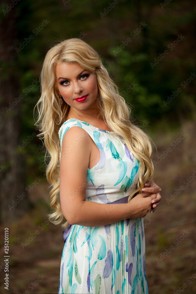 Young beautiful blonde woman in white dress with blue and green flowers posing in forest in summer Young beautiful blonde woman in white dress with blue and green flowers posing in forest in summer
