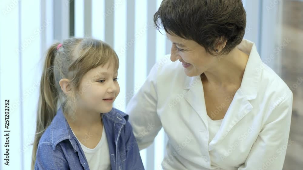 Tilt-up of smiling female doctor talking friendly with little patient sitting on medical examination couch Tilt-up of smiling female doctor talking friendly with little patient sitting on medical examination couch