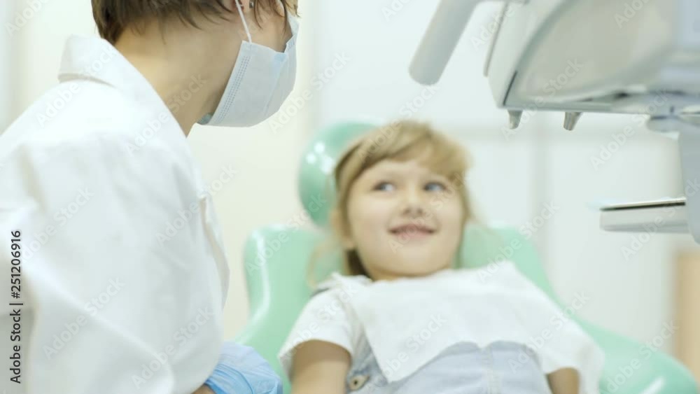 Anonymous female in mask and doctor uniform talking with sweet cheerful girl during dental examination Anonymous female in mask and doctor uniform talking with sweet cheerful girl during dental examination