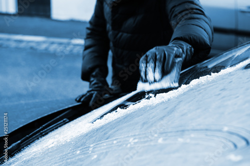 View from outside the car of woman cleaning car windshield from frost using specialized ice frost scraper and rubber squeegee car windscreen in cold winter blue toned image. photo