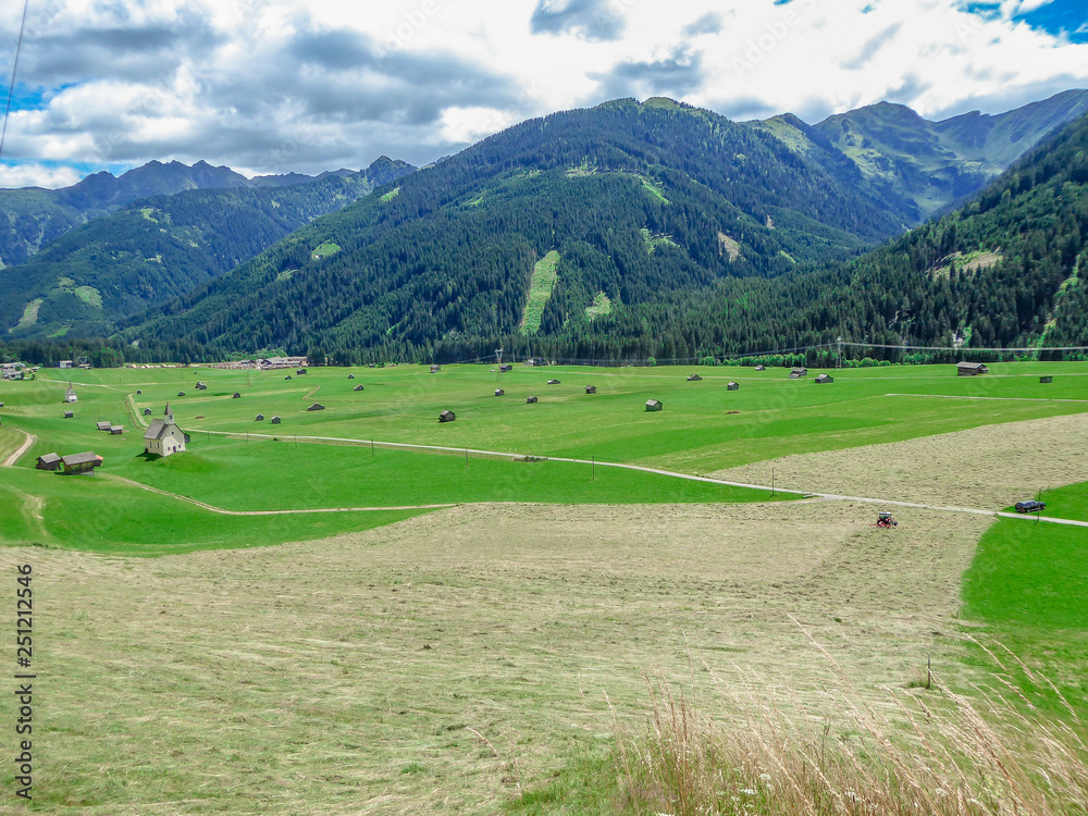 Spring came to the Alpine valley, turning the meadow into lush green color. Lots of small cottages and agricultural machinery on the grassland. High Alpine peeks around the valley. Small church.