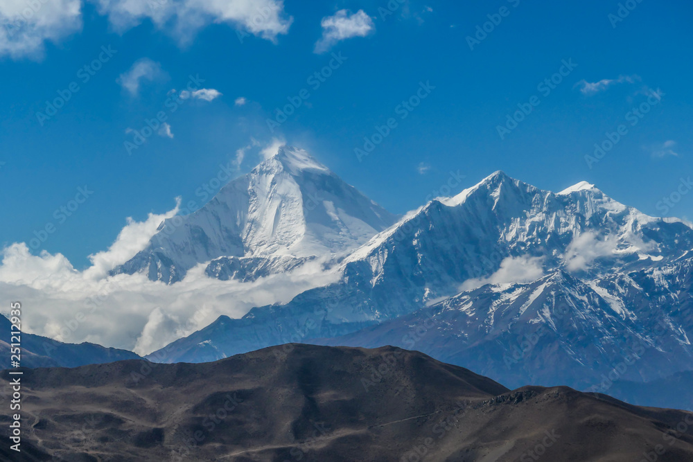 View on Himalayas from Annapurna Circuit Trek, Nepal. Clouds covered with clouds. Sharp slopes. Smaller mountain in front, covered in shadow.  Overcast but sunny. High altitude climbing.