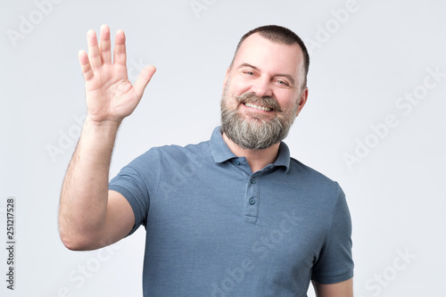 Fotografi Polite ymature man with beard dressed in denim shirt saying hi, waving with hand
