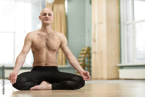 Horizontal shot of a shirtless young man practicing yoga indoors meditating in lotus position. Athletic male exercising at gym studio relaxing during meditation copy space.
