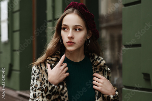 Outdoor close up fashion portrait of young beautiful fashionable lady wearing trendy animal, leopard print faux fur coat, beret, hoop earrings, walking in street of  European city