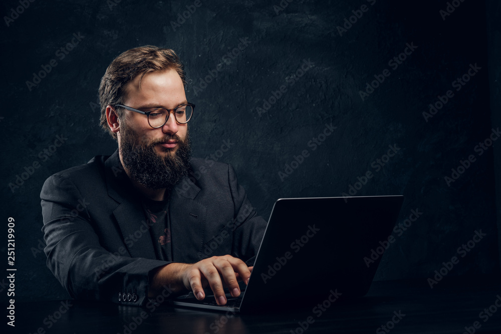 Bearded programmer in suit working on laptop sitting at the table in office against a dark wall Bearded programmer in suit working on laptop sitting at the table in office against a dark wall