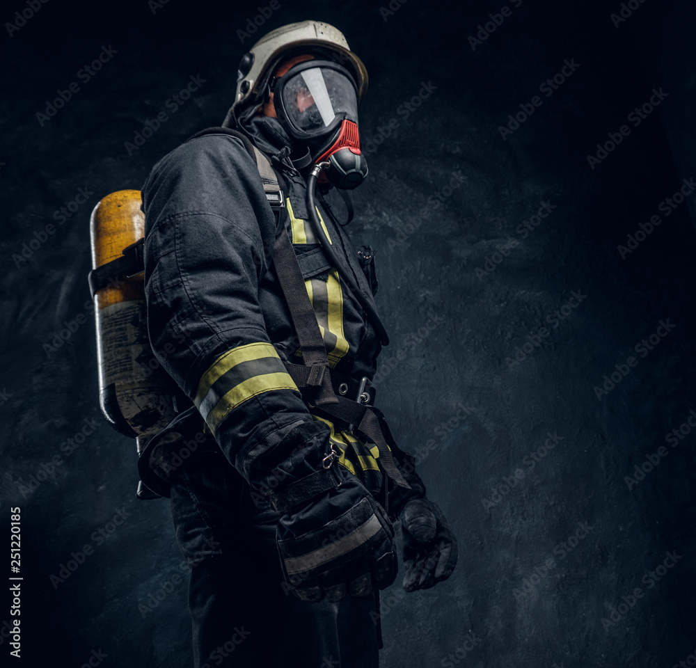 Naklejka premium Firefighter in safety helmet and oxygen mask wearing protective clothes. Studio photo against a dark textured wall