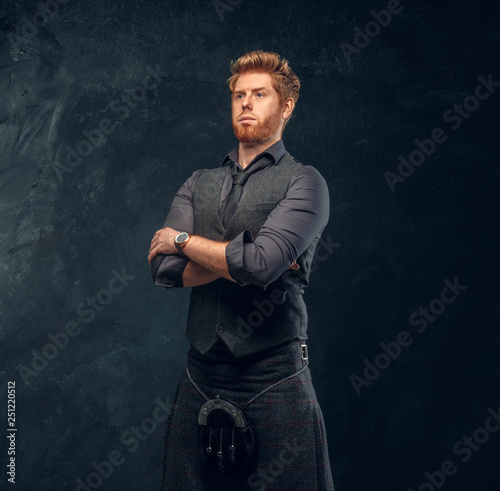 Handsome redhead man dressed in an elegant vest with tie and kilt posing with his arms crossed in studio against a dark textured wall