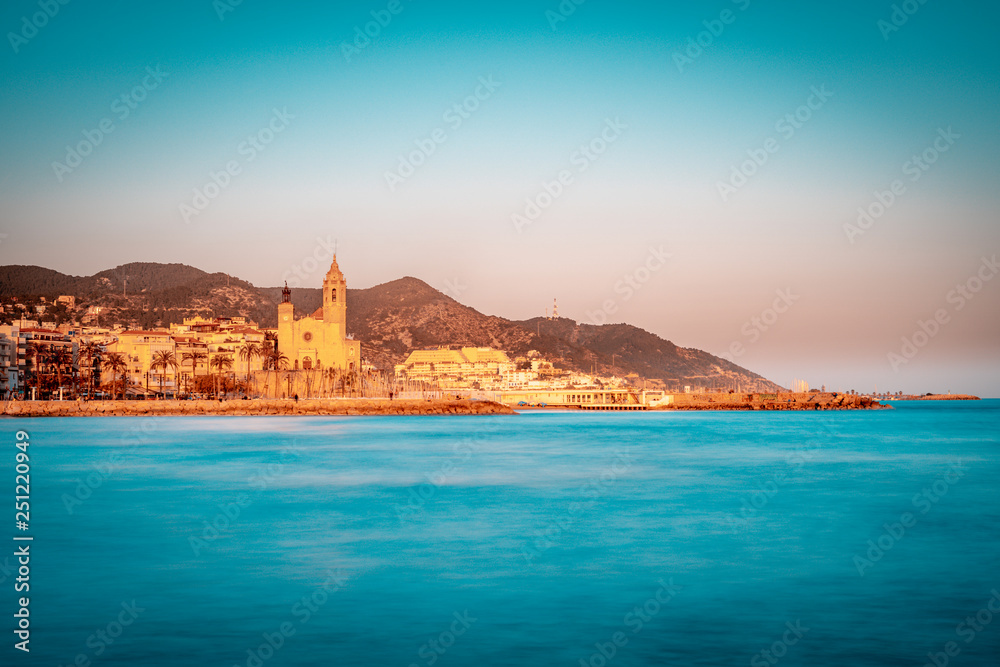 Orange and teal view of mediterranean town of Sitges, with the iconic Church of Saint Bartholomew and Santa Tecla, in the suburb area of Barcelona. Blurred sea waves by a slow shutter speed