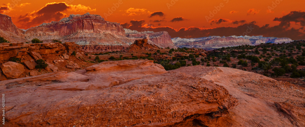 Sunset during golden hour in Southern Utah, sun warming red sandstone, cliffs, mountains, and mesa.  With orange sky and red clouds