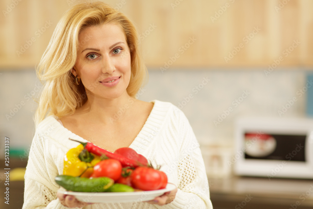 Beautiful happy healthy mature woman smiling to the camera posing at her kitchen with a plate full of delicious vegetables. Housewife preparing healthy diet meals at home