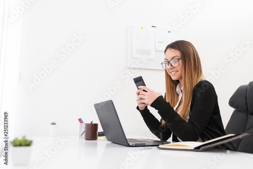 Photos Side view of joyful, beautiful economist checking her social medias on phone in front of the lap top, at the office
