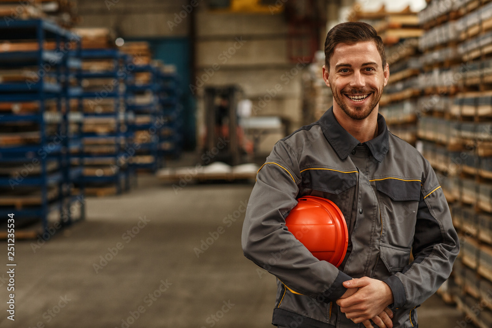 Shot of a handsome young bearded factory worker in uniform holding protective hardhat smiling joyfully to the camera posing at the warehouse of a metalworking company - obrazy, fototapety, plakaty 