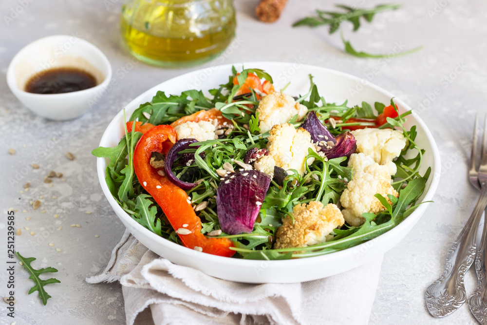 Salad with arugula, roasted vegetables (cauliflower, red bell pepper and onion) and seeds. Healthy food. Light grey concrete background. Copy space. Salad with arugula, roasted vegetables (cauliflower, red bell pepper and onion) and seeds. Healthy food. Light grey concrete background. Copy space.