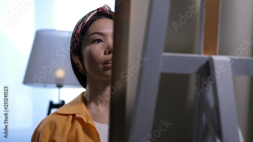 Focused face of charming asian woman painting on large easel in art studio. Close up view of young female painter expressing inspiration and imagination in artwork. Dolly shot photo