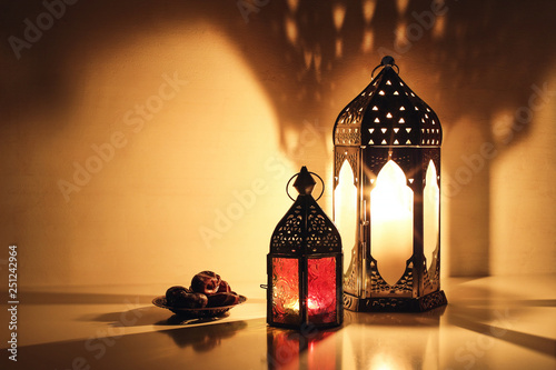 Ornamental Arabic lanterns with burning candles glowing at night. Plate with date fruit on the table. Festive greeting card, invitation for Muslim holy month Ramadan Kareem. Iftar dinner background.