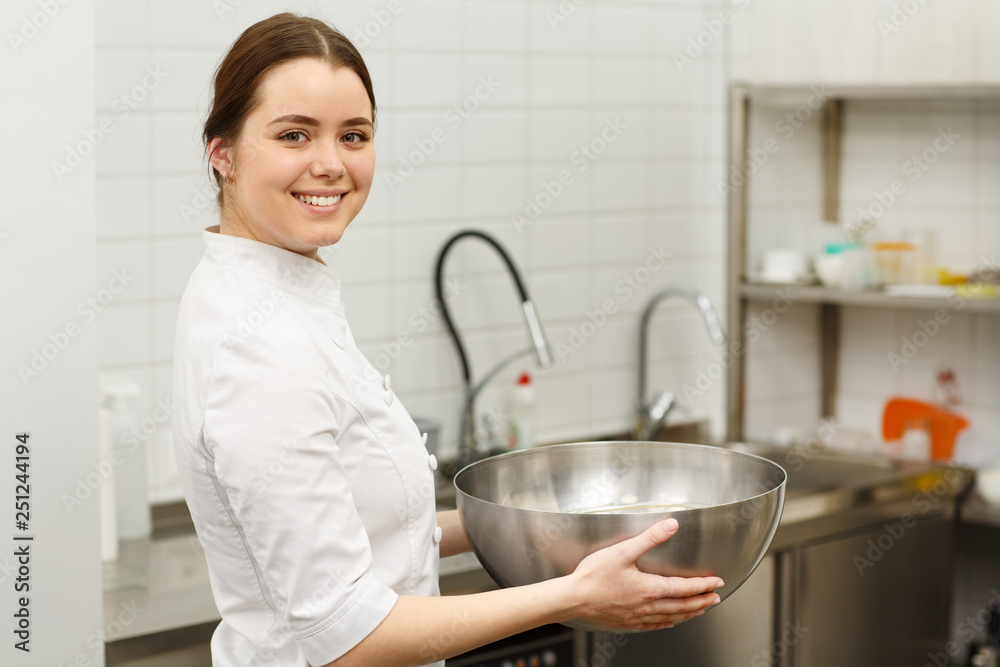 Smiling and beautiful girl holding big metal bowl with dough for some desserts. Pretty confectioner wearing white special uniform and standing in white modern confectionery. Smiling and beautiful girl holding big metal bowl with dough for some desserts. Pretty confectioner wearing white special uniform and standing in white modern confectionery.