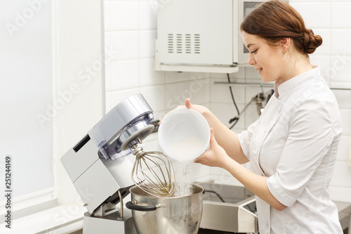 Pretty chef cooking cream for delicious  desserts and cakes using special modern kitchenware. Beautiful and handsome girl putting sugar in metal bowl of electric mixer.