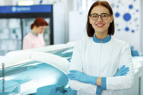 Front view of pretty lab assistant standing  looking at camera and smiling. Female lab technician in spectacles wearing special blue gloves and white uniform. Woman working in modern laboratory.