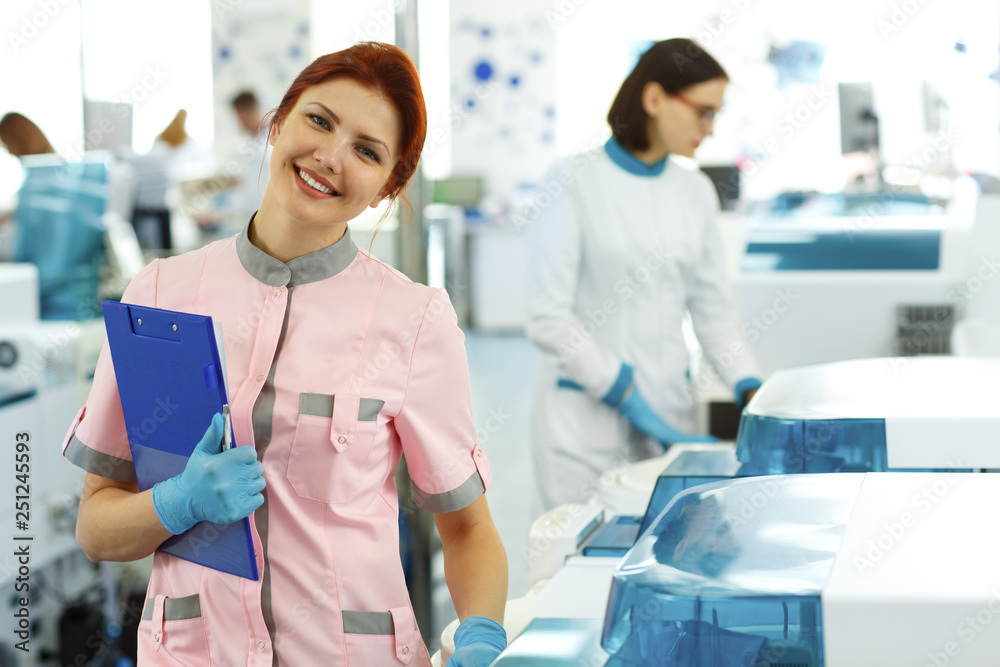 Female lab worker with ginger hair wearing pink uniform and holding dark blue folder. Woman smiling and posing. Medical workers and laboratory with modern equipment on background.