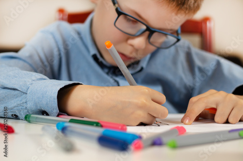 Chłopiec w niebieskiej koszuli i okularach siedzi przy białym biurku i w skupieniu rysuje cienkopisami na białej kartce papieru. Na pierwszym planie kolorowe flamastry. photo