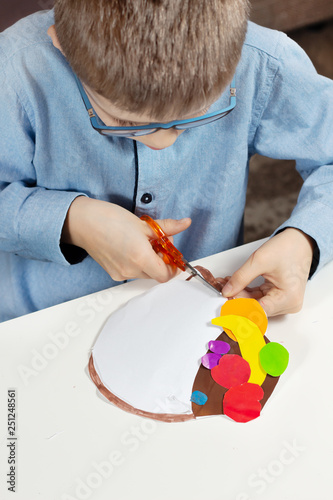 Chłopiec siedzi przy białym biurku i wycina nożyczkami kształt z kartki papieru. photo