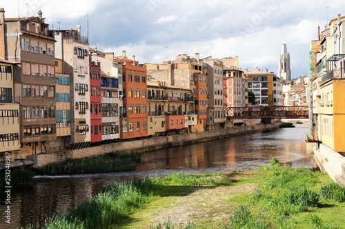 Colorful yellow, orange and red houses line the banks of the Onyar River in Girona, Catalonia, Spain with a bridge in the background © Caron