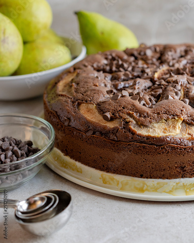 Close up view of left side of Chocolate Pear Cake with measuring spoons and chocolate chips in foreground.  Bowl of pears in background. photo