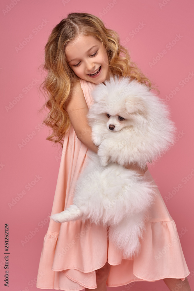 Little girl with a blond curly hair, in a pink dress plays with her dog Little girl with a blond curly hair, in a pink dress plays with her dog