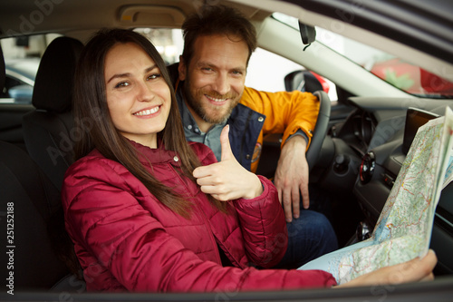 Pretty couple sitting in automobile cabin, smiling and looking at camera. Woman and man buying new car and planning trip. Beautiful woman holding map and showing thumb up. © Nestor