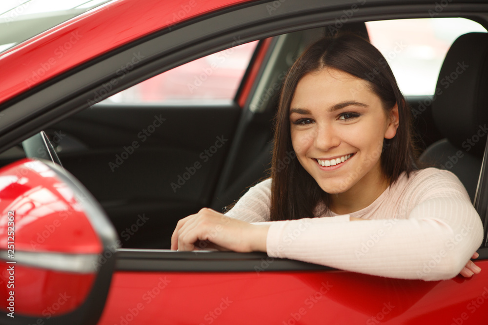 Pretty girl with brilliant smile looking at camera from window of red car. Happy young woman sitting in vehicle and enjoying comfortable automobile cabin. Purchase of new auto in car center.