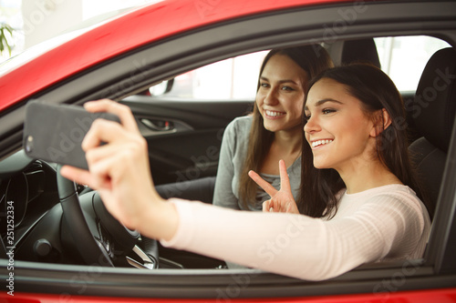 Two pretty girls making selfie in automobile cabin. Girls sitting inside, smiling, one girl holding phone. Female customers happy because of purchasing new auto in car center.