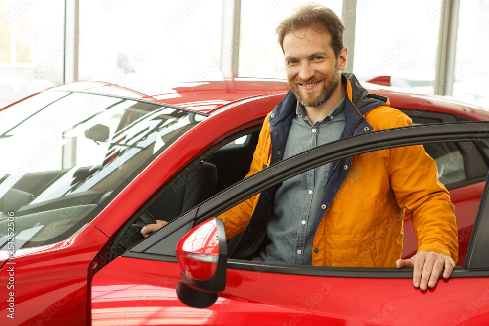 Handsome red haired male customer standing with opened door, near vehicle, smiling and looking at camera. Man enjoying comfortable red car, want buy new automobile in modern car dealership.