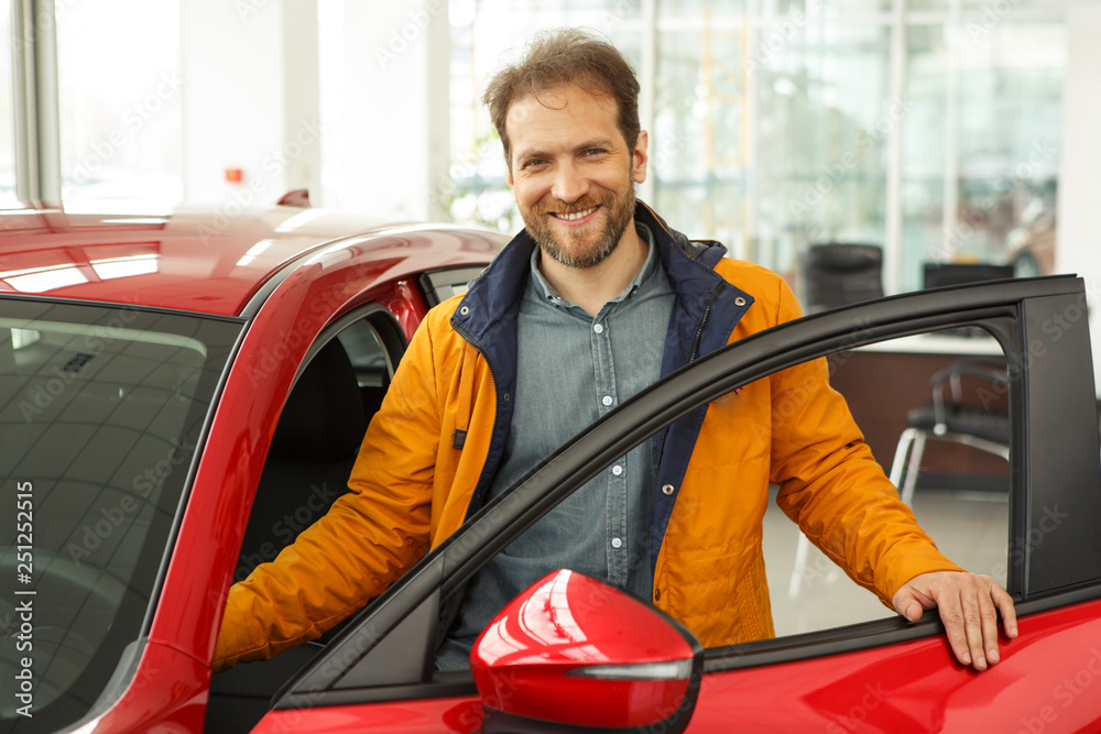 View of positivity man in yellow jacket standing near red car with opened door and smiling at camera. Handsome male customer posing in modern car center while choosing new automobile.